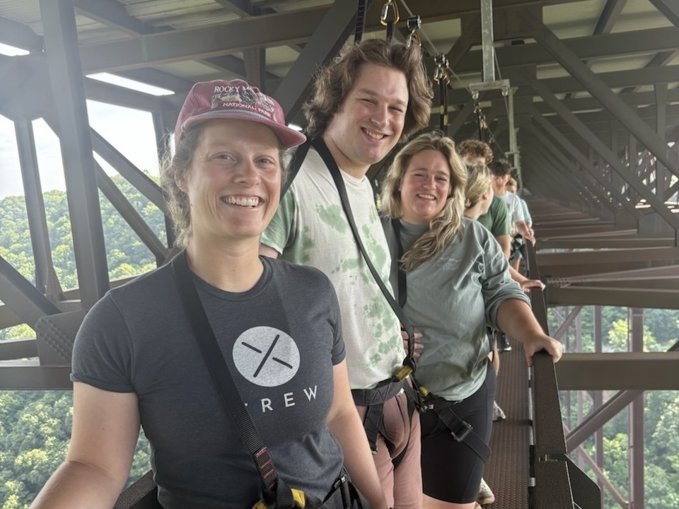 Three people with blonde-ish hair stand on a bridge walkway, strapped in by harnesses.