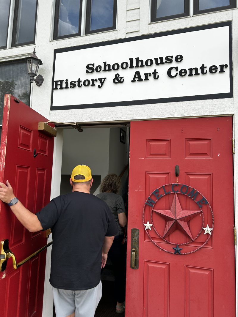 A man wearing a yellow hat walks through a bright red door, and a sign above it reads: Schoolhouse, History and Art Center