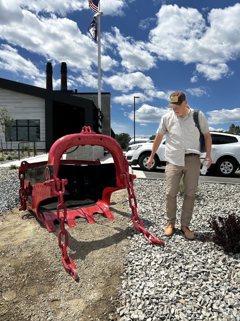 A man wearing a baseball hat and a short-sleeved button down shirt walks past a bright red coal bucket in front of a new building
