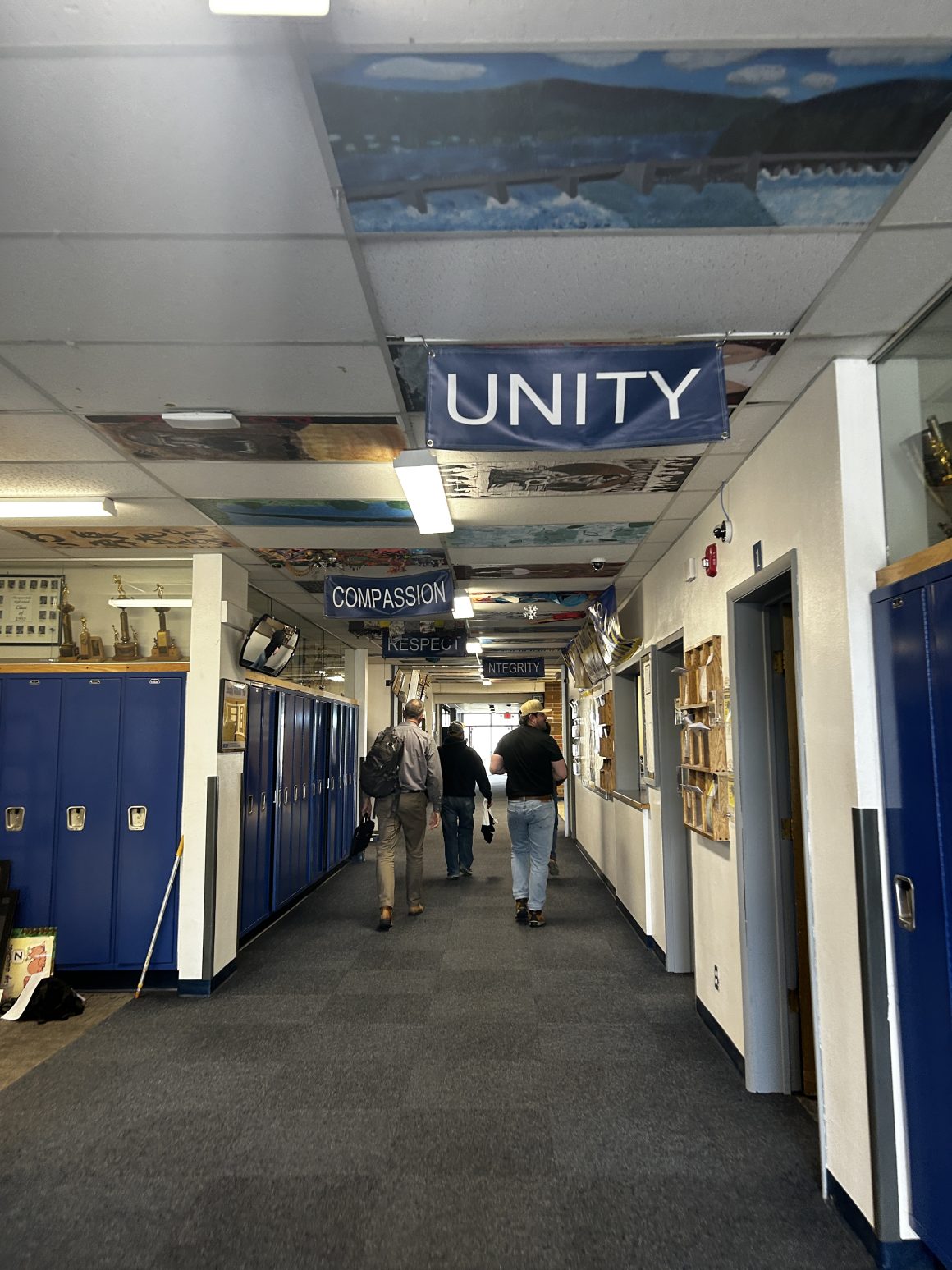 A group of people walk down a high school hallway with motivational blue banners hanging from the ceiling, reading words like "Unity"