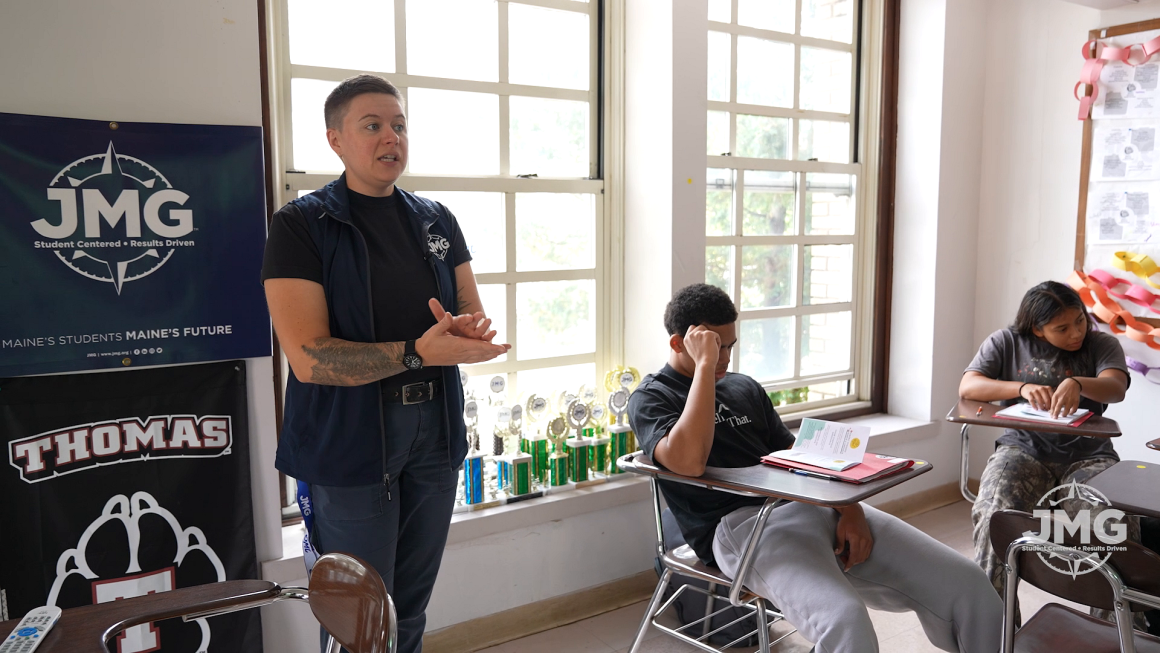An educator with cropped hair and a tattoo wears a vest with the letters JMG on it while standing in front of a classroom with students at desks.