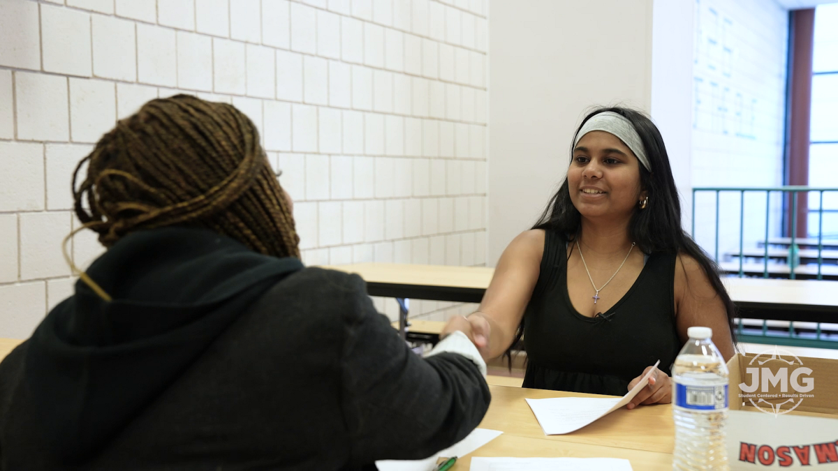Two young women shake hands as they sit across from each other in a mock interview