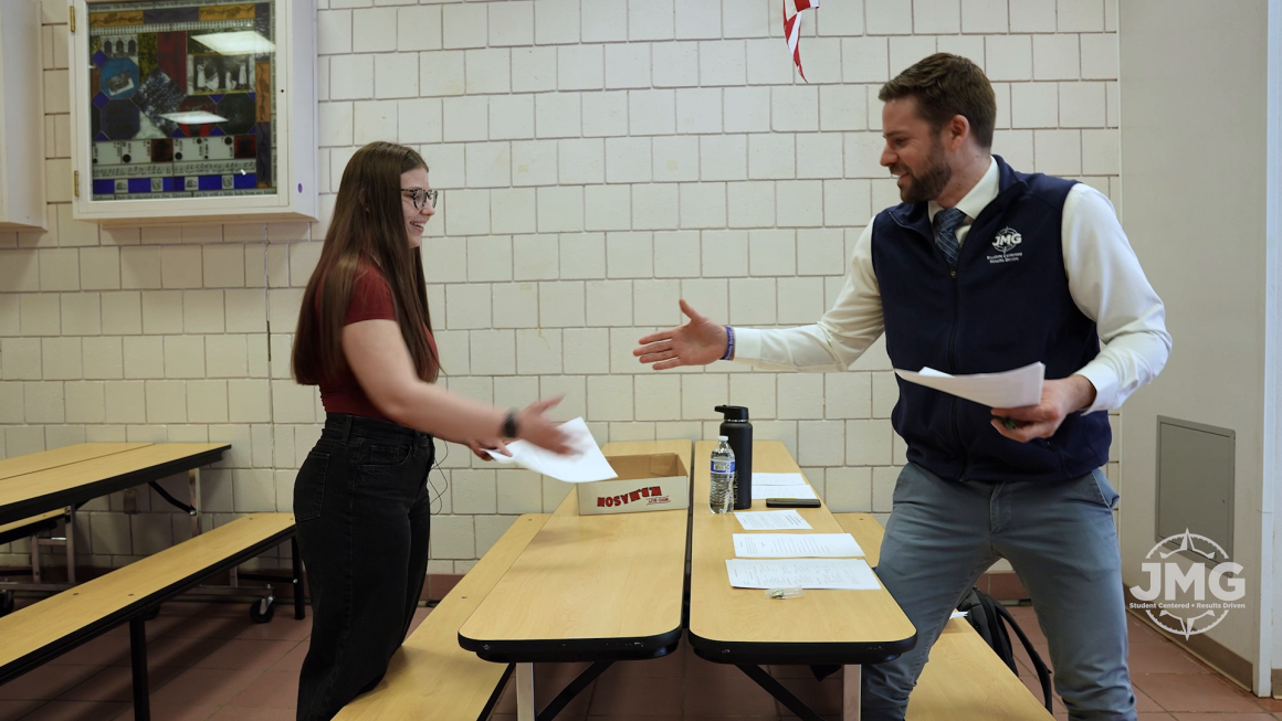 A young woman with long hair and glasses stands across from her instructor at a cafeteria table, preparing to shake his hand. Both smile.