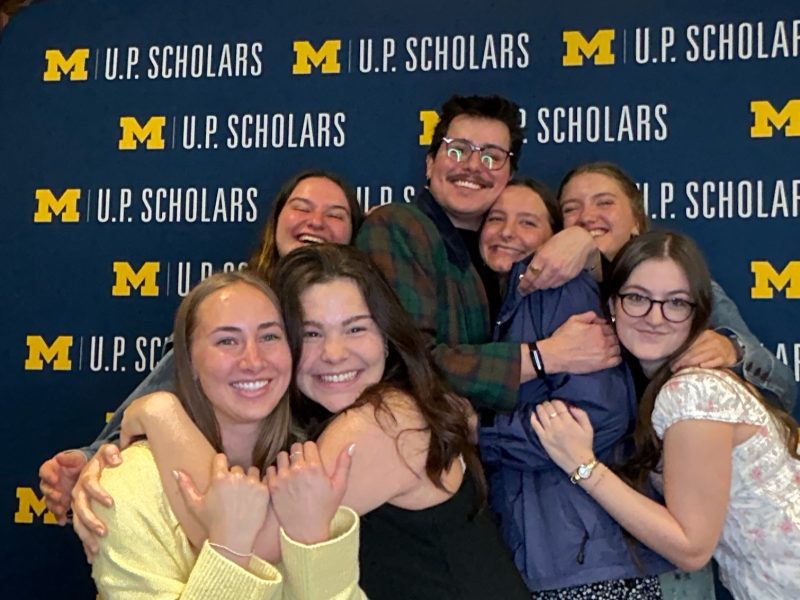 a group of seven students embracing in front of a Michigan U.P. Scholars backdrop, smiling